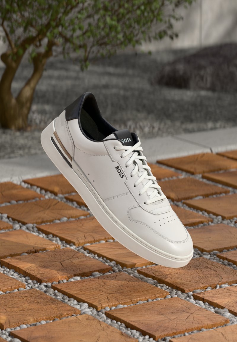 White leather sneaker with black heel accent, lace-up design, perforated toe, and branding. Elevated above a patterned stone surface.