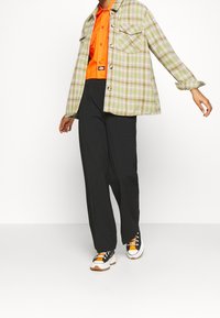 Green and beige plaid shirt jacket over an orange button-up, paired with black trousers and white sneakers featuring orange and black accents.