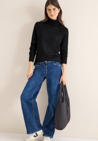Schwarzer Strickpullover mit Rollkragen, weite blaue Jeans und weiße Sneakers mit schwarzen Akzenten; hält eine graue, gepolsterte Tragetasche.