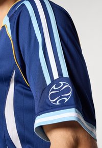 Αθλητική μπλούζα ναυτικού τόνου με οριζόντιες ρίγες σε ανοιχτό μπλε και λευκό. Περιλαμβάνει κεντημένο λογότυπο στο μανίκι και ριμπ μανσέτες.