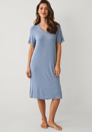 Kvinde stående barfodet iført en lyseblå knælængde kjole med korte ærmer omkranset af blonder og en V-udskæring.