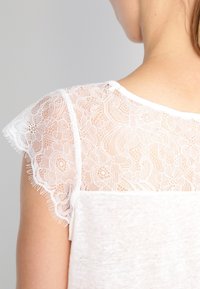 Bílý top s krajkovým designem rukávů a květinovým vzorem, jemnou texturou a kulatým výstřihem. Krajka má vlnité okraje.