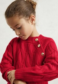 Crvena pletena puloverica s teksturiranim uzorkom čavla, rebrastim izrezom i tri zlatne dugmadi na ramenu. Udoban i prilagođen dizajn.