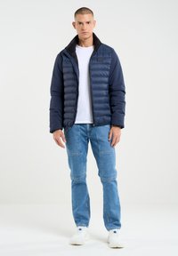 Jūras spēku puffer jaka ar quiltētu priekšpusi, melnu apkakli, apvienota ar gaiši ziliem džinsiem un baltām sporta kurpēm, ar priekšējo rāvējslēdzēju.