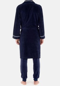 Accappatoio blu navy in pile con cintura, con polsini a righe bianche. Abbinato a pantaloni aderenti coordinati e piedi nudi visibili.