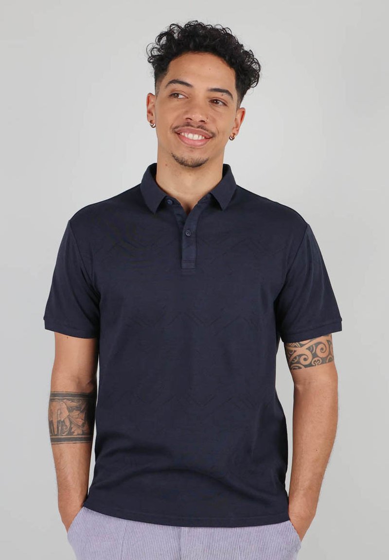 Polo en bleu marine en tissu doux et texturé avec col et fente à trois boutons ; présente des manches courtes et des fentes latérales.