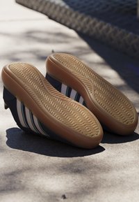 Par de zapatos negros con rayas blancas y verdes que muestran suelas de goma marrón claro con patrón de espiga en el suelo de cemento.