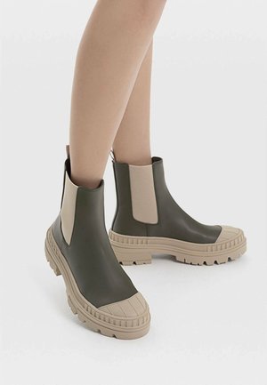 Bottines Chelsea en caoutchouc vert avec des panneaux élastiques beige sur les côtés, semelle texturée et capuchon de protection renforcé. Design imposant avec une apparence robuste.