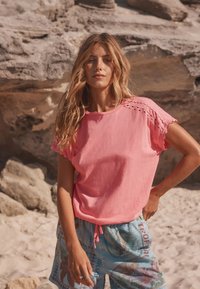 Rózsaszín rövid ujjú ing hullámos szegéllyel, könnyű anyagból készült. Világoskék, trópusi mintás rövidnadrággal párosítva.