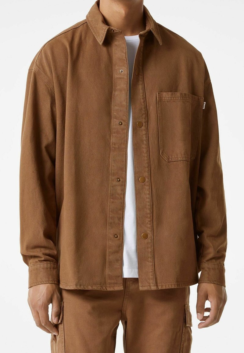 Brązowa koszula zapinana na guziki, wykonana z wytrzymałego materiału, z kieszonką na piersi, zatrzaskami i klasycznym kołnierzykiem. Nosi się ją na białym T-shircie.