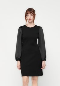 Black dress with fitted bodice, round neckline, and black satin puff sleeves. Features a knee-length hem with a side slit for mobility.