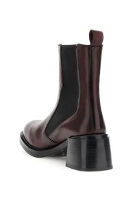 Botină de gleznă din piele maro închis, cu panou elastic lateral negru, brâu de prindere și toc gros negru, prezentată din unghiul din spate.