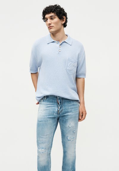 Νέος άνδρας με σγουρά μαλλιά, φοράει ανοιχτό μπλε κοντομάνικο πόλο με τσέπη και ξεφτισμένο μπλε τζιν, στέκεται με τα χέρια πίσω από την πλάτη.
