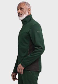 Grüne Fleecejacke mit hohem Kragen, Reißverschlusstaschen, strukturiertem Stoff und schwarzen Akzenten an den Seiten.