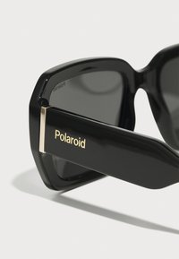 Occhiali da sole neri con montatura spessa, dotati di un accento dorato sulla tempia. Testo "Polaroid" in oro. Le lenti sono scure e riflettenti.