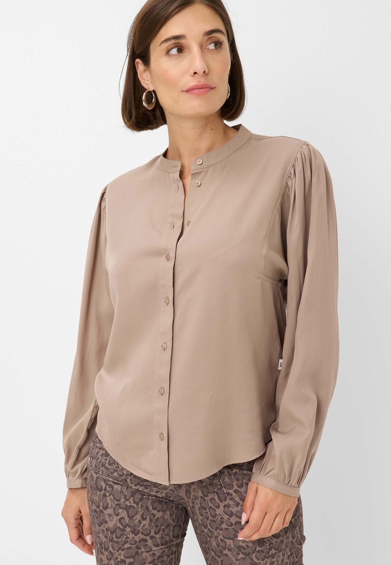 Blusa beige a maniche lunghe con colletto a fascia, chiusura frontale a bottoni e maniche arricciate. Tessuto morbido e scivoloso. Abbinata a pantaloni con stampa animale.
