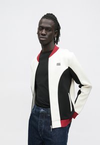 Chaqueta bomber blanca y negra con detalles en rojo, que cuenta con cierre de cremallera, logo y un diseño curvo contrastante. Se lleva sobre una camisa negra y vaqueros.