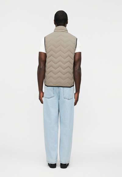 Άνδρας φορώντας μπεζ γιλέκο χωρίς μανίκια με καπιτονέ, λευκό κοντομάνικο πουκάμισο, ανοιχτό μπλε τζιν και μαύρα παπούτσια, γυρισμένος προς τα πίσω σε απλό φόντο.