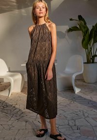 Robe midi en dentelle marron avec un col halter, présentant une silhouette fluide et des motifs en vagues complexes. Le mannequin porte des sandales noires.