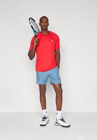 Punane niiskust eemaldav spordipoliis, millel on väike logo. Heledat sinist lühikesed püksid, valged sokid logoga ning valged tennisepoed tumedate aktsentidega.