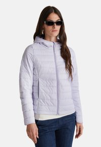 Světle fialová péřová bunda s kapucí, s předním zipem a elastickými manžetami, nošená přes bílý svetr a džíny.