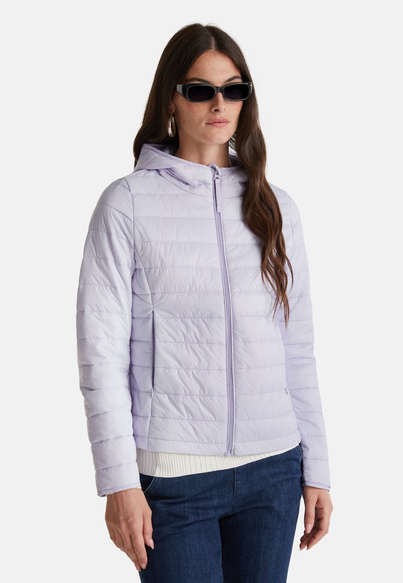 Světle fialová péřová bunda s kapucí, s předním zipem a elastickými manžetami, nošená přes bílý svetr a džíny.