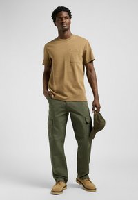T-shirt di cotone beige con taschino sul petto, abbinato a pantaloni cargo verdi. L'outfit include un cappellino da baseball verde scuro e scarpe beige.