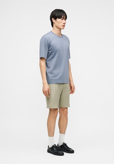 Νέος άνδρας που στέκεται πλάγια φορώντας ανοιχτό μπλε μπλουζάκι, μπεζ σορτς, λευκές κάλτσες και μαύρα αθλητικά παπούτσια σε απλό λευκό φόντο.