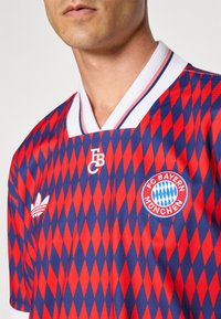 Czerwona i niebieska koszulka FC Bayern Monachium z wzorem w romby, białym kołnierzykiem i wyszywanym logo. Wykonana z gładkiego, lekkiego materiału.