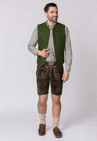 Gilet di lana verde sopra una camicia a quadri, abbinato a pantaloni corti di pelle marrone con cuciture decorative, bottoni in ottone e calze al ginocchio.