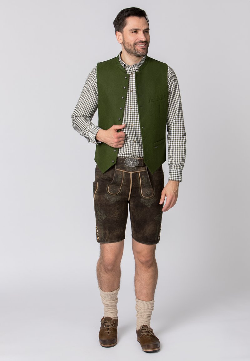 Gilet di lana verde sopra una camicia a quadri, abbinato a pantaloni corti di pelle marrone con cuciture decorative, bottoni in ottone e calze al ginocchio.