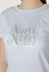T-shirt bianco in cotone con logo di "Armani Exchange" in glitter argento sul petto. Collo rotondo e maniche corte.