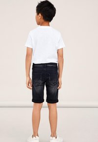 Pantalones cortos de mezclilla oscura con dobladillos doblados, que cuentan con dos bolsillos traseros y un acabado suave, combinados con una camiseta blanca lisa y zapatillas blancas.