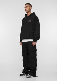 Felpa nera oversize con tasca frontale e logo, abbinata a pantaloni cargo con più tasche, tutto in nero, completato da sneakers con motivo.