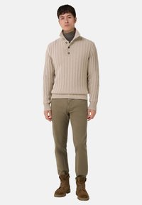 Beżowy prążkowany sweter z wysokim kołnierzem, trzema guzikami i długimi rękawami, zestawiony z oliwkowozielonymi spodniami i brązowymi zamszowymi butami.