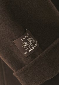 Barna gyapjúszövet texturált felülettel. Fekete címke fehér szöveggel és három medve illusztrációval. A címke tartalmazza a márkát és az évet.