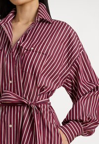 Rødt og hvitt stripete oversized skjorte med spiss krage, knappestolpe foran, to fremre lommer og beltedetalj i midjen.