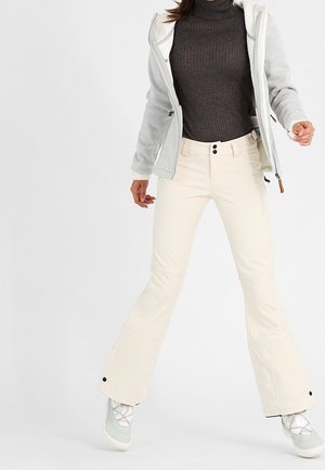 Hellgraue Kapuzenjacke aus strukturiertem Material, gerippter dunkler Rollkragenpullover und cremefarbene Hose. Auffällige Merkmale: Auslaufende Hosenbeine und graue Schuhe.