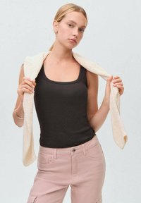 Zwarte geribbelde tanktop met een vierkante halslijn, gecombineerd met lichtroze broek. Een licht crèmekleurige gebreide trui hangt over de schouders.