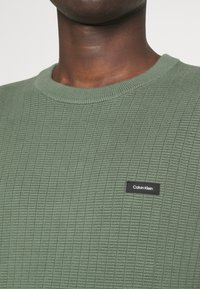 Maglione verde a maglia con un motivo testurizzato, caratterizzato da un collo rotondo a coste e una piccola etichetta nera con il logo di Calvin Klein sul davanti.