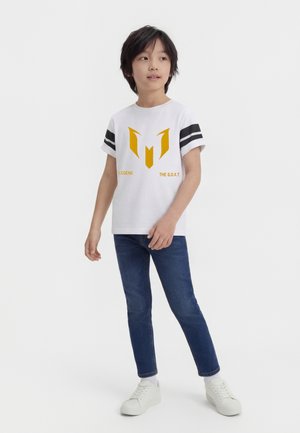 Ragazzo con capelli di media lunghezza che indossa una maglietta bianca con logo e testo gialli, jeans blu e sneakers bianche, in piedi e che guarda di lato.