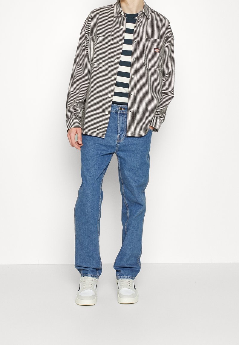 Homme portant une chemise boutonnée rayée noir et blanc par-dessus un T-shirt rayé noir et blanc, un jean bleu et des baskets blanches, debout devant un fond blanc.
