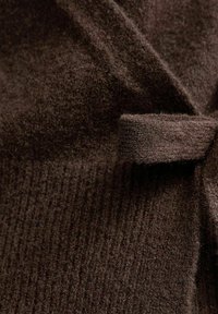 Tessuto marrone con una superficie morbida e testurizzata, caratterizzata da sezioni a coste e un dettaglio annodato. Il materiale appare caldo e denso.