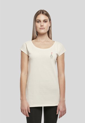 Jeune femme aux cheveux blonds et raides porte un t-shirt crème à manches courtes avec un petit motif de main noire et cœur rouge sur la poitrine.