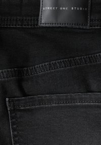 Tessuto in denim nero con due tasche frontali, cuciture in evidenza e un'etichetta in pelle impressa con "STREET ONE STUDIO" nella parte superiore.