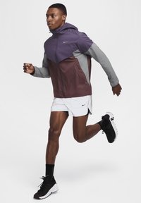 Chaqueta de running Nike en púrpura, gris y granate; material ligero, con cremallera en la parte delantera y diseño con capucha. Lleva pantalones cortos blancos y zapatillas negras.