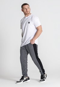 Weißes T-Shirt mit kleinem schwarzem Logo, gemusterte schwarze Jogginghose mit weißen Akzenten und schwarze Sneaker mit weißen Details.