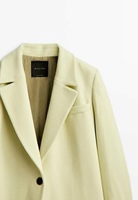 Manteau vert clair en mélange de laine avec un revers à cran, une fermeture à un bouton et une poche poitrine. Texture douce et lignes épurées.