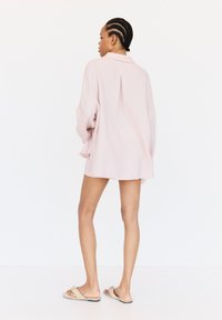 Lichtroze oversized shirt met lange mouwen en een rugplooitje, gedragen over shorts. De geweven stof complementeert het ontspannen ontwerp.