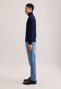 Pull à col roulé bleu marine en tissu tricoté doux, associé à un jean droit bleu clair et des chaussures à lacets noires.
